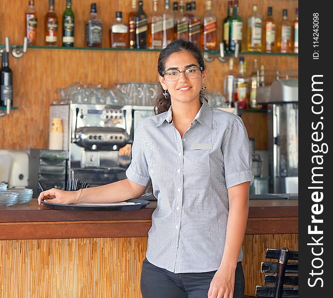 The waitress is standing near the restaurant bar of the hotel. A waitress with a tray is waiting for the order. Shelves with bottles of alcohol in the background. The concept of service. The waitress is standing near the restaurant bar of the hotel. A waitress with a tray is waiting for the order. Shelves with bottles of alcohol in the background. The concept of service.