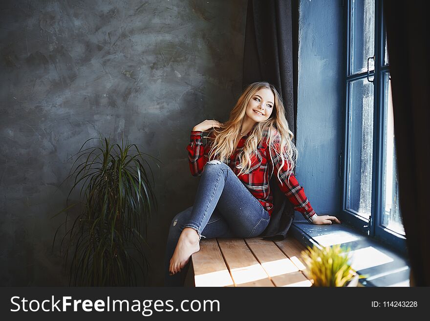 Happy and smiling beautiful blonde girl in jeans and red plaid shirt, sits and posing near the window and looks outside