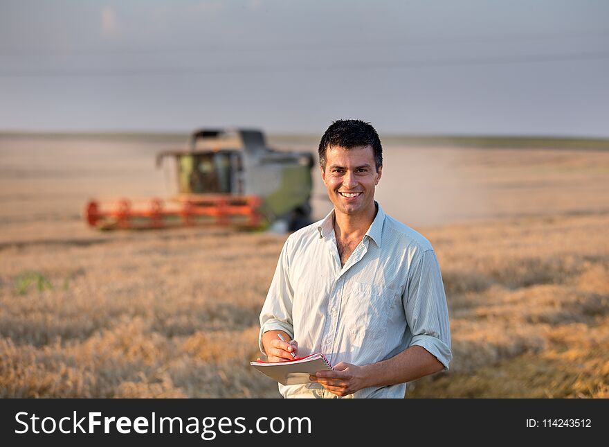 Happy young farmer engineer with notebook standing on wheat field while combine harvester working in background. Happy young farmer engineer with notebook standing on wheat field while combine harvester working in background