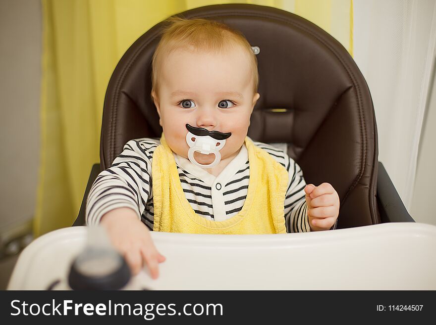 Little funny boy sitting in a chair for feeding with childish nipple in the form of a mustache. Indoor