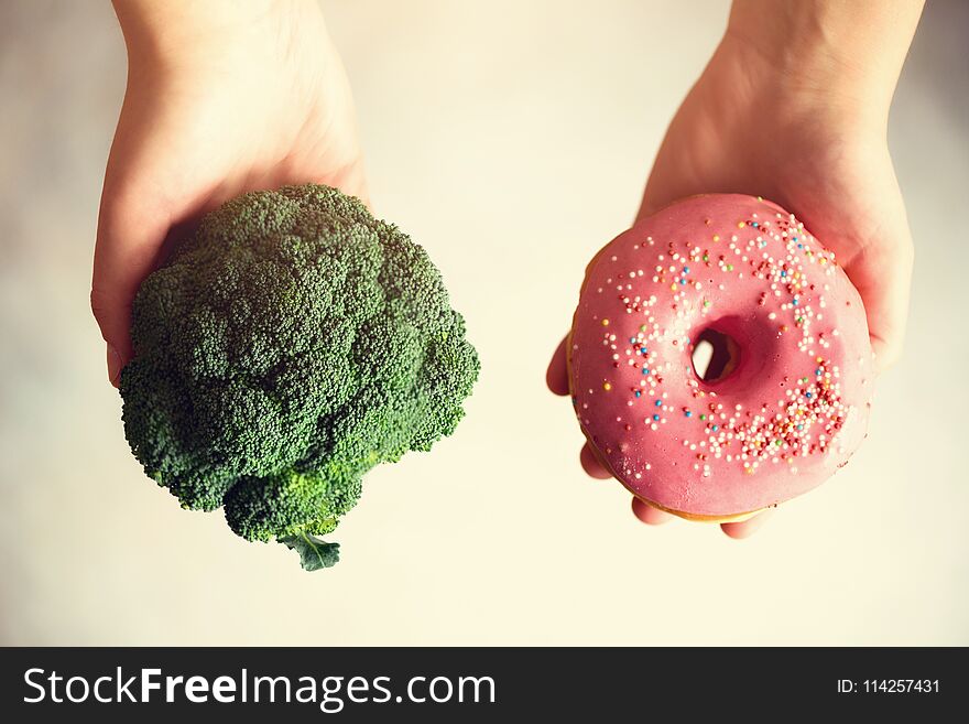 Young woman in white T-shirt choosing between broccoli or junk food, donut. Healthy clean detox eating concept. Vegetarian, vegan, raw concept. Copy space.