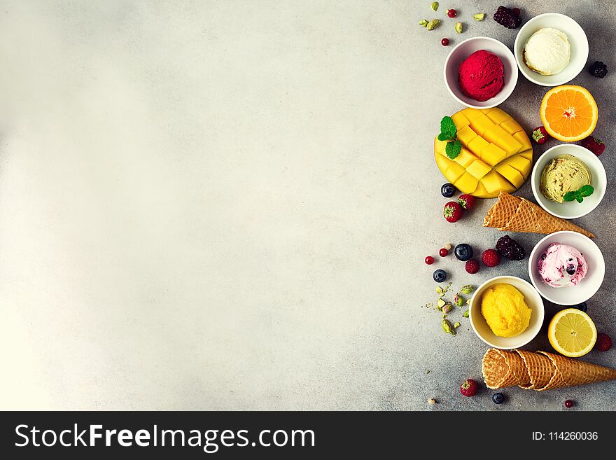 Ice cream balls in bowls, waffle cones, berries, orange, mango, pistachio on grey concrete background. Colorful