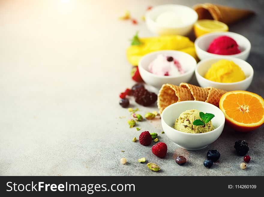 Ice cream balls in bowls, waffle cones, berries, orange, mango, pistachio on grey concrete background. Colorful