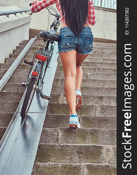 Back View Of A Slim Female Dressed A Pink Flannel Shirt And Denim Shorts, Climbs The Stairs With A Bike.