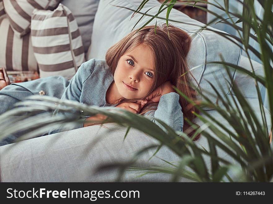 Portrait of a cute little girl with long brown hair and piercing glance, looking at a camera, lying on a sofa at home alone.