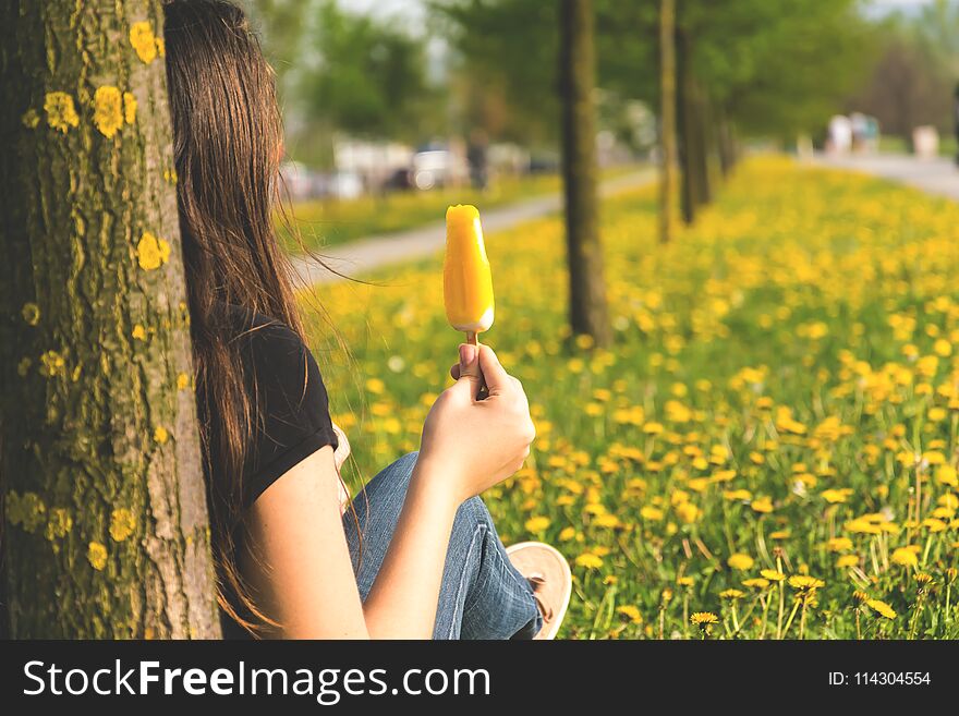 Girl sits leaning on a tree and eating ice cream surrounded with yellow dandelion flowers. Spring time. Girl sits leaning on a tree and eating ice cream surrounded with yellow dandelion flowers. Spring time.
