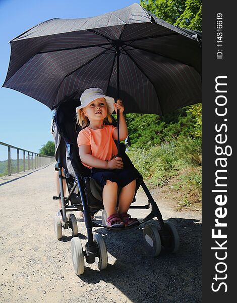 Little girl sitting in stroller. Child protected from sun by big umbrella. Kid Heatstroke prevention on sunny day in summer