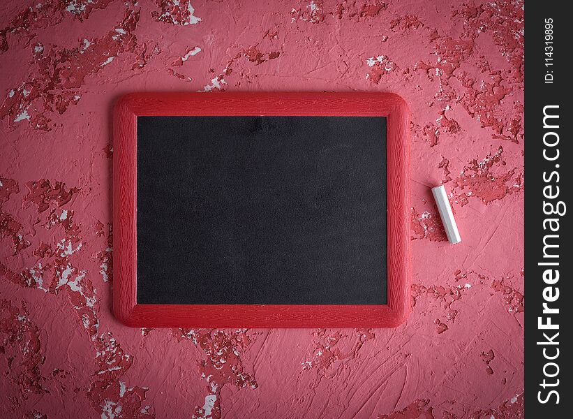 An empty black chalk board in a red frame and a piece of chalk on a worn red surface, close up