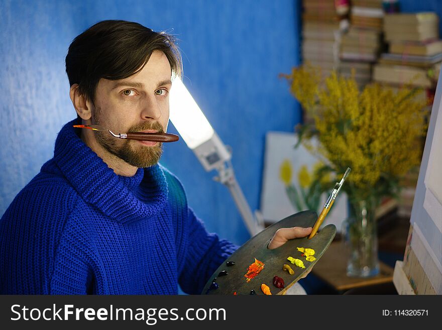 Artist holds a palette in his mouth. He holds palette with oil paints and brushes. Canvas and flowers in background. Artist holds a palette in his mouth. He holds palette with oil paints and brushes. Canvas and flowers in background