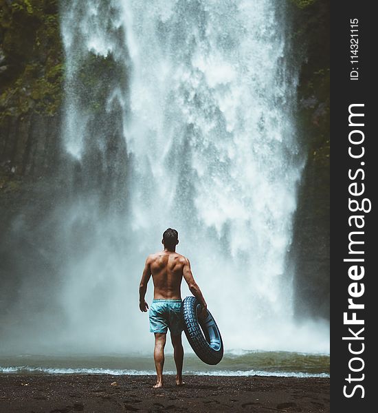 Man Wearing Blue Shorts Holding Vehicle Tire Facing Waterfalls