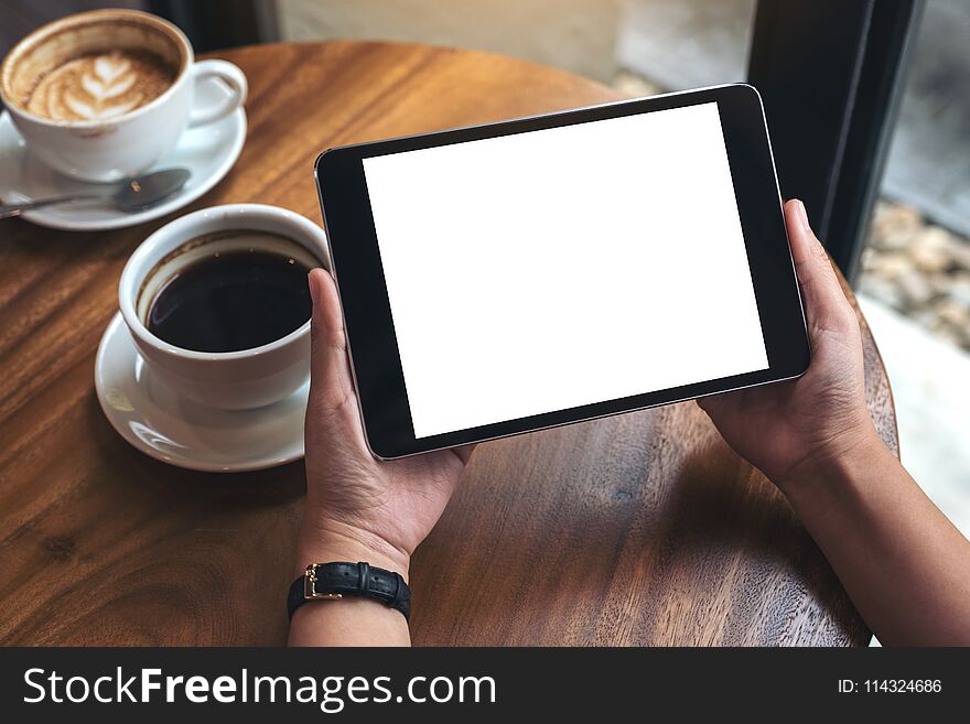 Hands holding black tablet pc with white blank screen and coffee cups on table