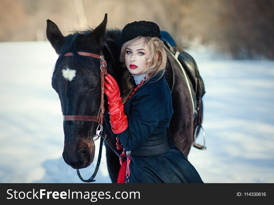 Blond woman in red dress with long black coat with dark horse on the background of winter landscape. Blond woman in red dress with long black coat with dark horse on the background of winter landscape