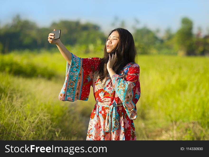 Young beautiful and happy Asian Chinese tourist woman on her 20s with colorful dress taking selfie pic with mobile phone camera on tropical green grass field background in holidays and travel concept