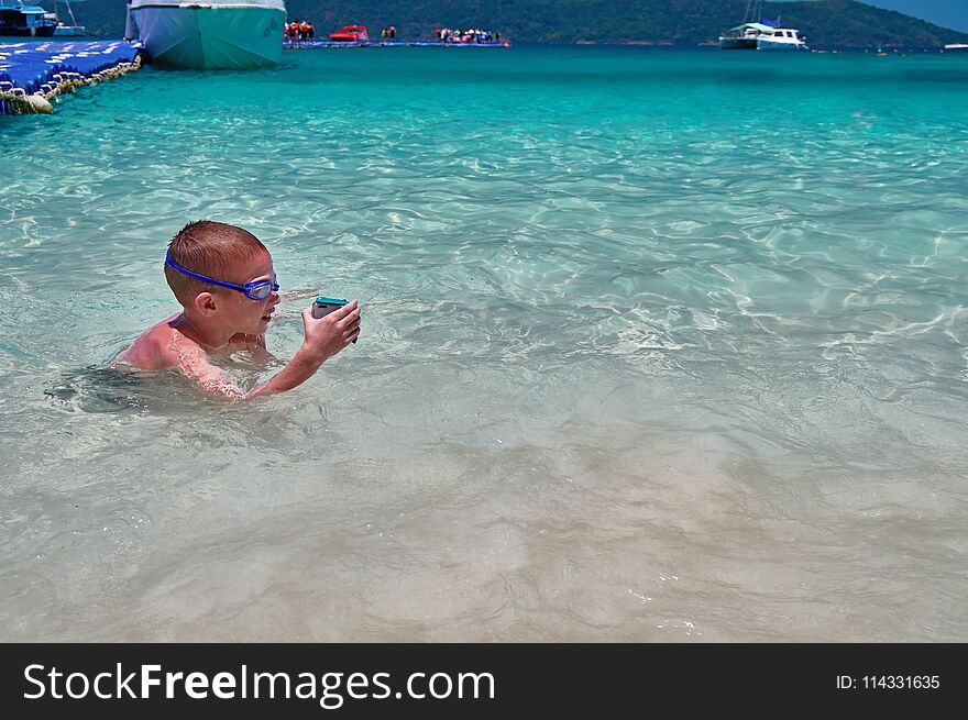 Little boy in swimming glasses plays in turquoise water with action camera in protective box. Child swims in waters tropical sea.