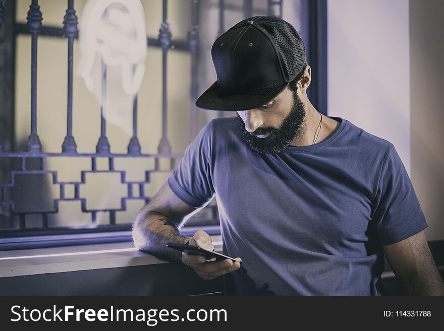 Young Bearded Man Sitting In Cafe And Using Smartphone For Texting Message. Horizontal. Blurred Background.