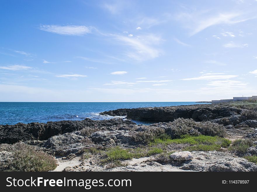 Wavy Sea Beside Rock Formations Near Sea Shore With Grasses Under Blue Cloudy Sky at Daytime