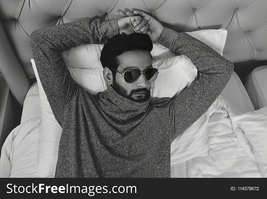 Grayscale Photo of Man in Long-sleeved Wearing Sunglasses Lying on Bed With Both Hand on Head