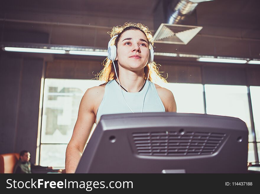 Theme is sport and music. A beautiful inflated Caucasian woman running in a gym on a treadmill. On her head are big white headphones, the girl listens to music during a cardio workout for weight loss. Theme is sport and music. A beautiful inflated Caucasian woman running in a gym on a treadmill. On her head are big white headphones, the girl listens to music during a cardio workout for weight loss.