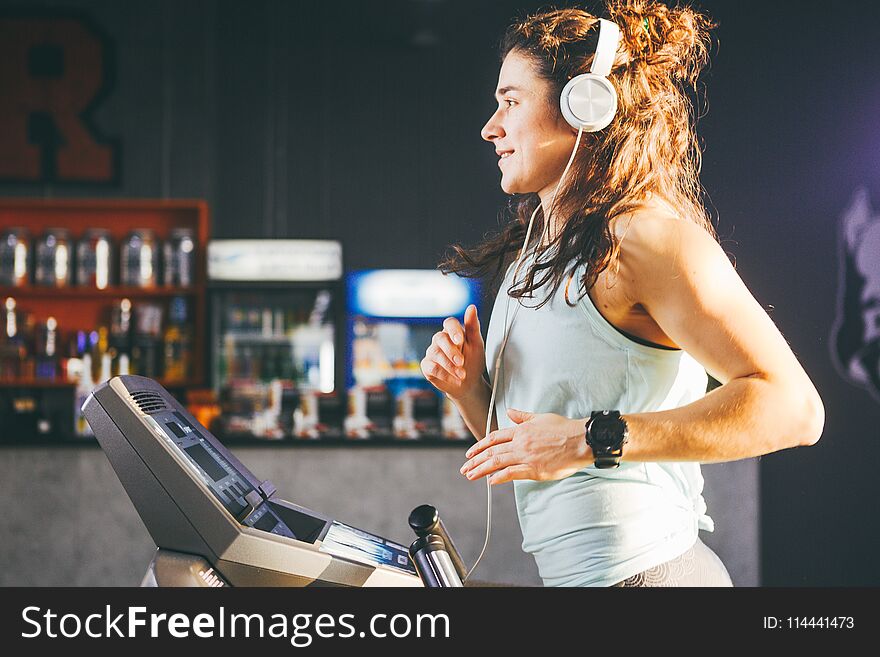 Theme is sport and music. A beautiful inflated woman runs in the gym on a treadmill. On her head are big white headphones, the girl listens to music during a cardio workout for weight loss.