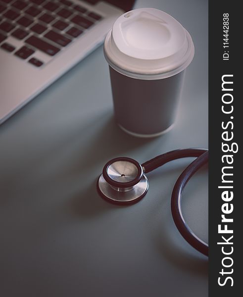 Gray and Black Stethoscope Beside Disposable Cup and Macbook Pro