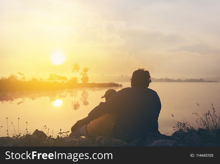 Two People On Grass Beside Body Of Water During Golden Hour