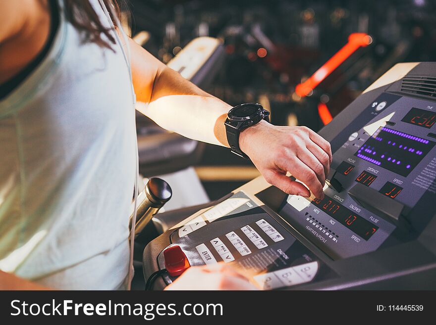 Close-up of a young Caucasian woman`s hand in the gym uses a sports watch, a black pulse wrist on the back of a treadmill in a spotlight in sunny weather.