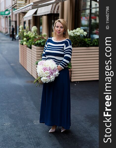 Young blonde woman in blue dress with peonies bouquet outdoors at city street