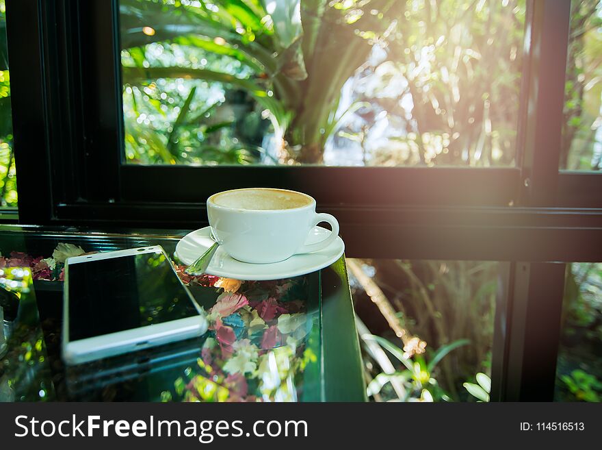 Picture of a white cup of coffee by the window. There is a smartphone next to it.At cafe