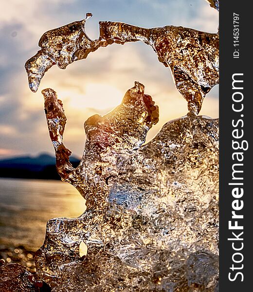 Shiny cold ice, bright icicles on frozen lake shore. Warm sun rays of sunset. Reflection in deep clefts