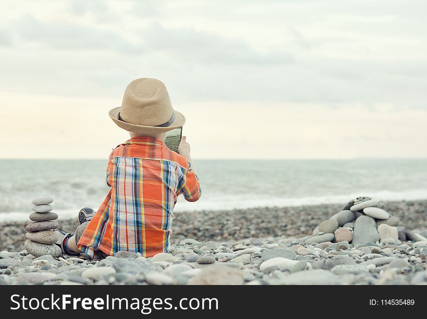 Little boy takes pictures on the smart phone. Sits on a pebble beach.