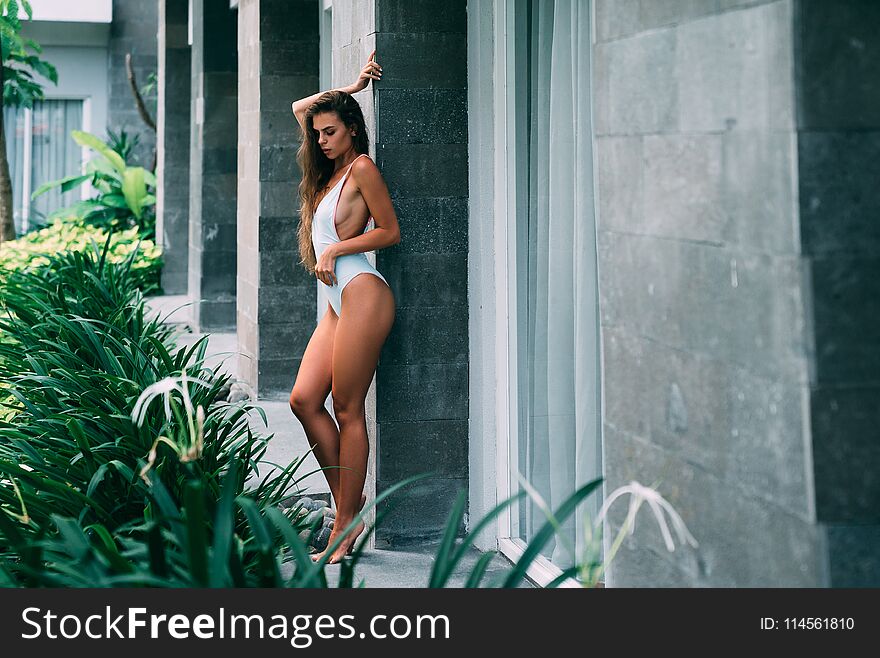 Beautiful brunette girl in white swimsuit posing in tropical location with green trees. Young sports model in bikini