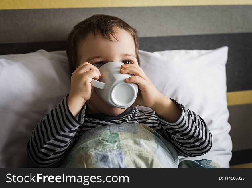 Unhealthy little boy lying on bed on pillow under blanket drinking medicine syrup in cup. Unhealthy little boy lying on bed on pillow under blanket drinking medicine syrup in cup