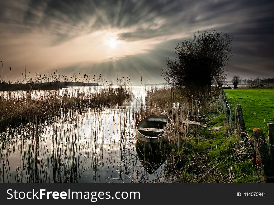 Old landscape with vintage rowing boat in a reed collar with atmospheric and intense sunset. Old pollard willows and drawn cloudy skies with sunbeams scorch the Dutch landscape with views over the lake. Old landscape with vintage rowing boat in a reed collar with atmospheric and intense sunset. Old pollard willows and drawn cloudy skies with sunbeams scorch the Dutch landscape with views over the lake.