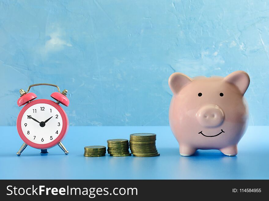 On a dark blue background photographed piggy bank, next to her a small pink alarm clock and three stacks of coins. Preservation of savings. A bright picture.