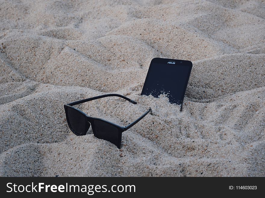 Black Wayfarer-style Sunglasses Beside Black Asus Android Smartphone on Brown Sand