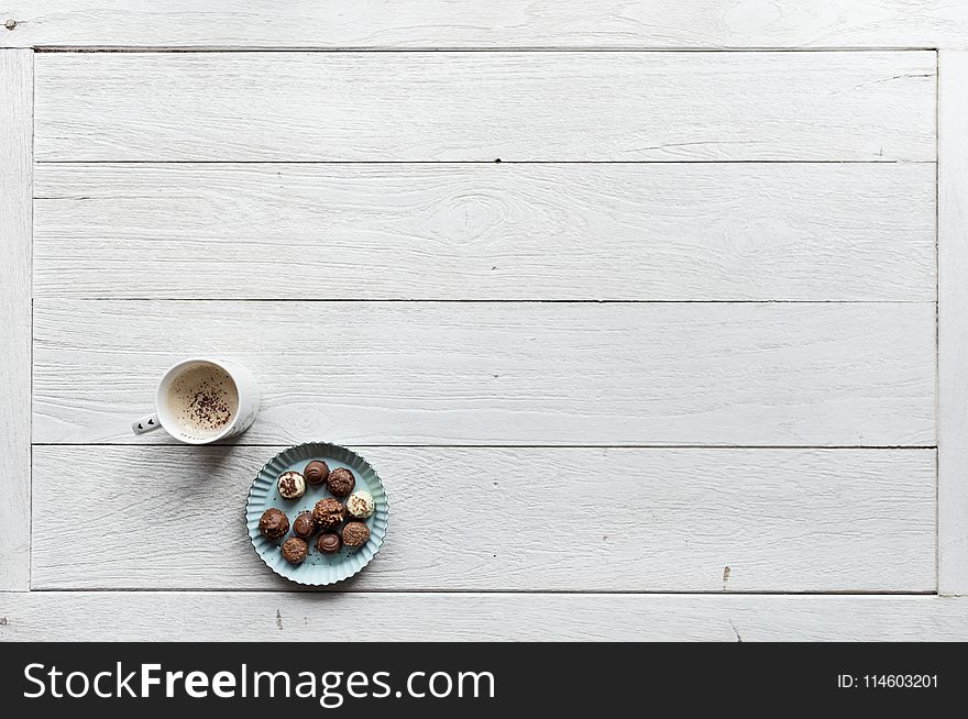 White Mug Filled With Coffee Beside Baked Pastries on Paper Plate All on Top of White Wood Surface