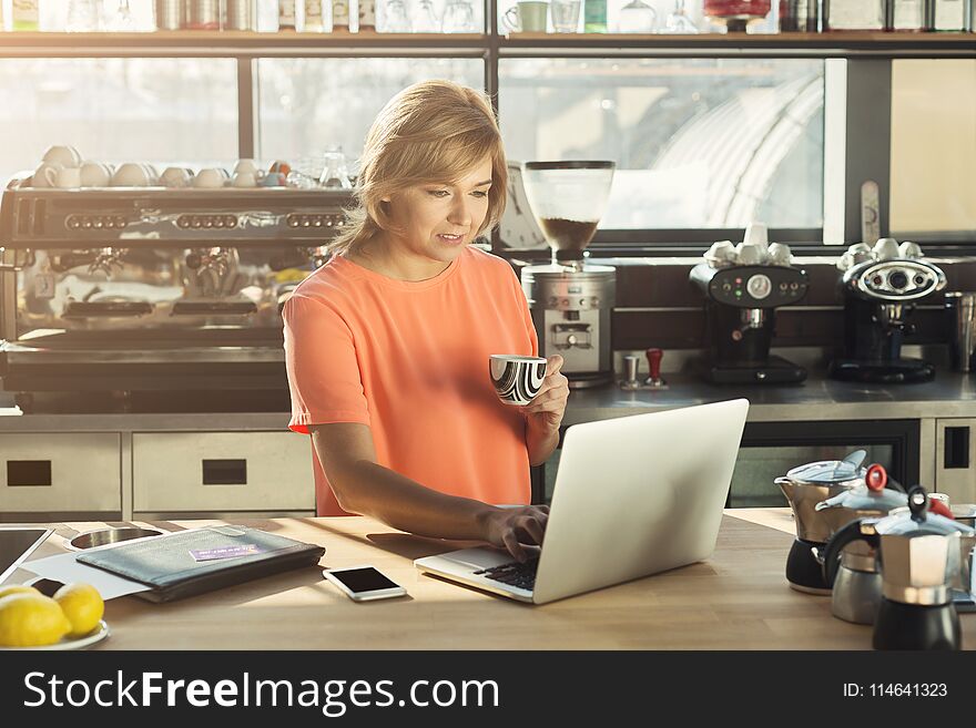 Middle-aged woman barista or manager working on laptop, making an order online and drinking coffee at modern cafeteria. Work, lifestyle, cafe, restaurant concept. Middle-aged woman barista or manager working on laptop, making an order online and drinking coffee at modern cafeteria. Work, lifestyle, cafe, restaurant concept