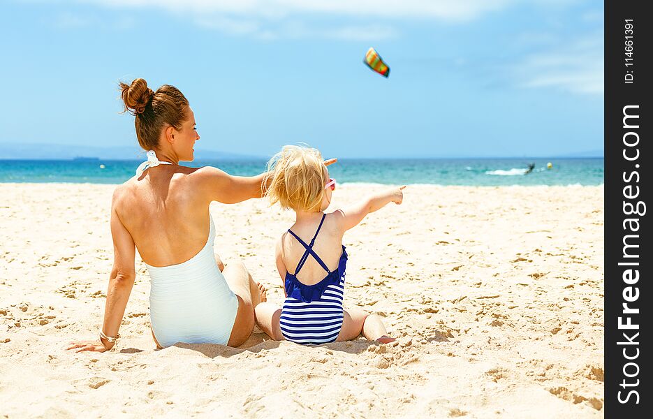 Family fun on white sand. Seen from behind modern mother and child in beachwear on the beach pointing at something. Family fun on white sand. Seen from behind modern mother and child in beachwear on the beach pointing at something