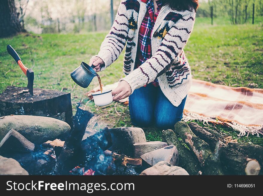 A girl is drinking coffee by the bonfire. A woman is preparing a morning coffee on fire. The brunette pours the drink into the cup. Camping in the forest. Picnic in nature. A girl is drinking coffee by the bonfire. A woman is preparing a morning coffee on fire. The brunette pours the drink into the cup. Camping in the forest. Picnic in nature.