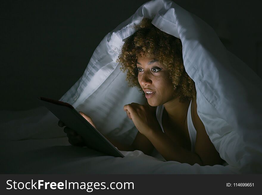 Charming young woman relaxing on bed in nighttime lying under cozy blanket and watching tablet excitedly in dark. Charming young woman relaxing on bed in nighttime lying under cozy blanket and watching tablet excitedly in dark.