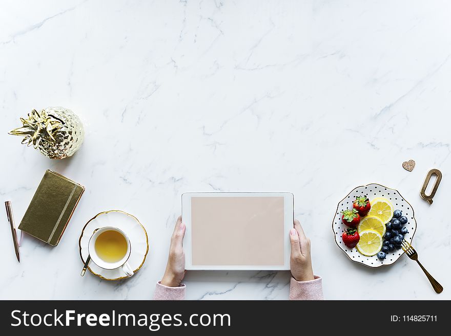 Person Holding White Tablet Computer Beside Teacup and Saucer of Blueberries