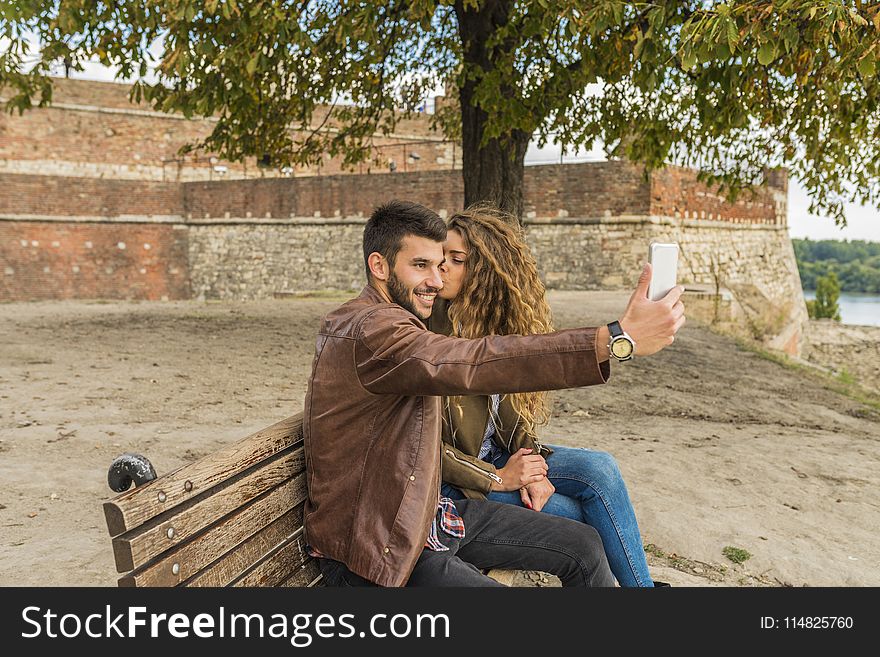 Woman And Man Sitting On Brown Wooden Bench Kissing