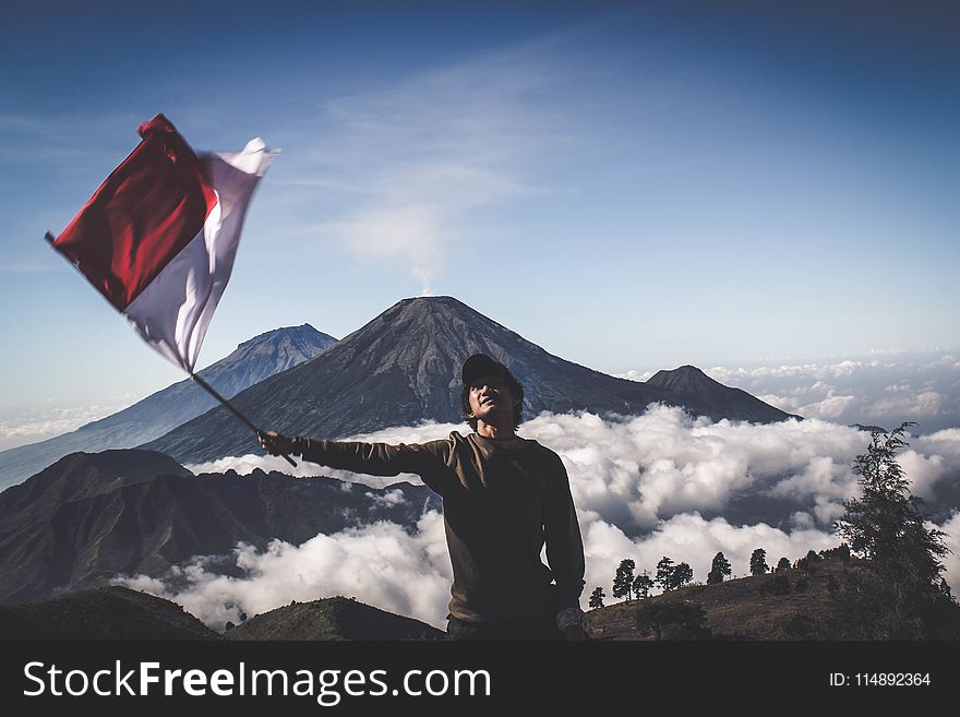 Man Wearing Black Crew-neck Sweater Holding White and Red Flag Standing Near Mountain Under Blue and White Sky