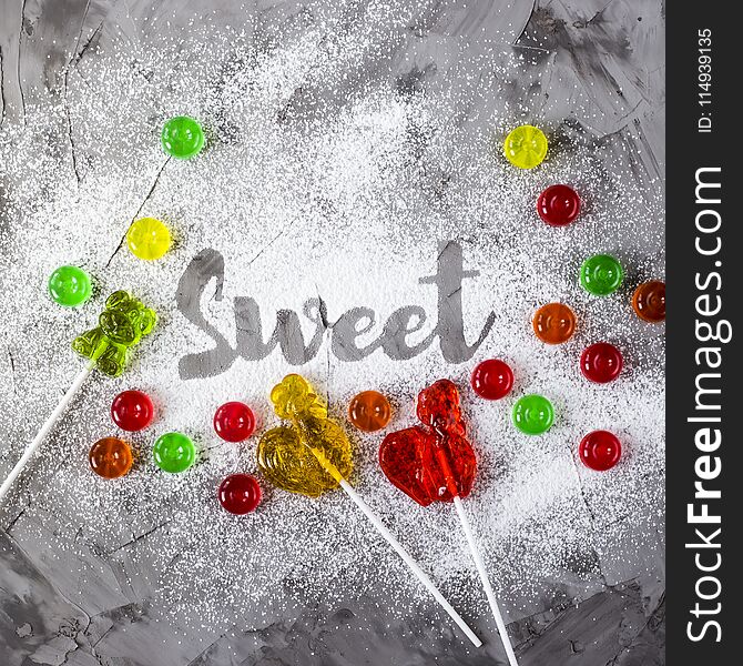 The word sweet is written on a gray concrete background with the help of powdered sugar next to multi-colored sweets and retro candies in the form of cockerels. Top view, flat lay. The word sweet is written on a gray concrete background with the help of powdered sugar next to multi-colored sweets and retro candies in the form of cockerels. Top view, flat lay
