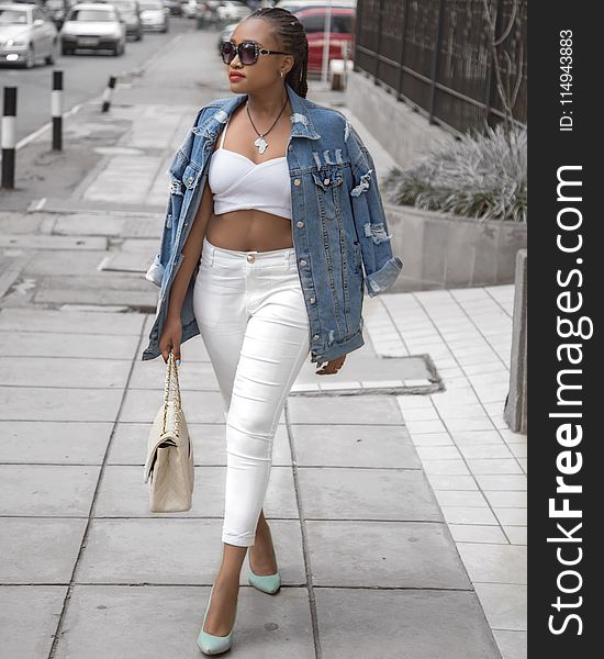 Photo of Woman Wearing White Crop Top and Denim Jacket Walking in Pavement Holding Bag