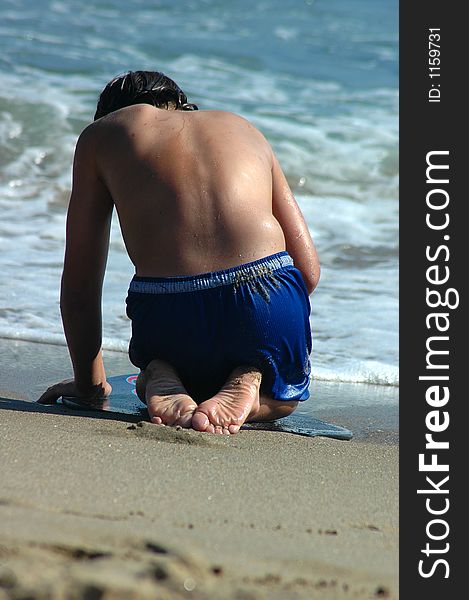 A boy is sitting on the skim board in the beach and 
looking for something in the sand. A boy is sitting on the skim board in the beach and 
looking for something in the sand