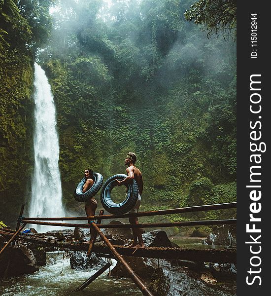 Two Person Carrying Black Inflatable Pool Float On Brown Wooden Bridge Near Waterfalls