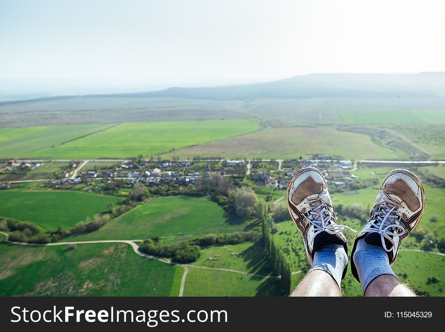 Male legs in white sneakers lit by sun over cliff against backdrop of countryside and green meadows.