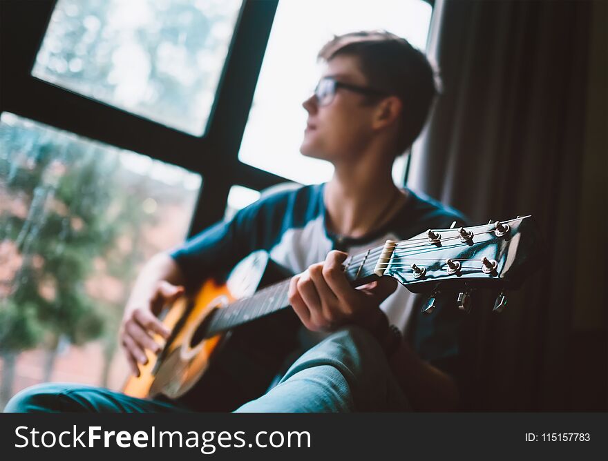 Young man plays on guitar sitting near the window