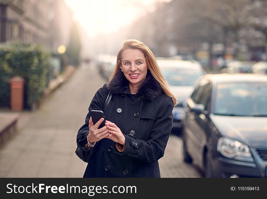 Attractive stylish woman with long blond hair using her mobile in an urban street in a busy town smiling as she reads a text message